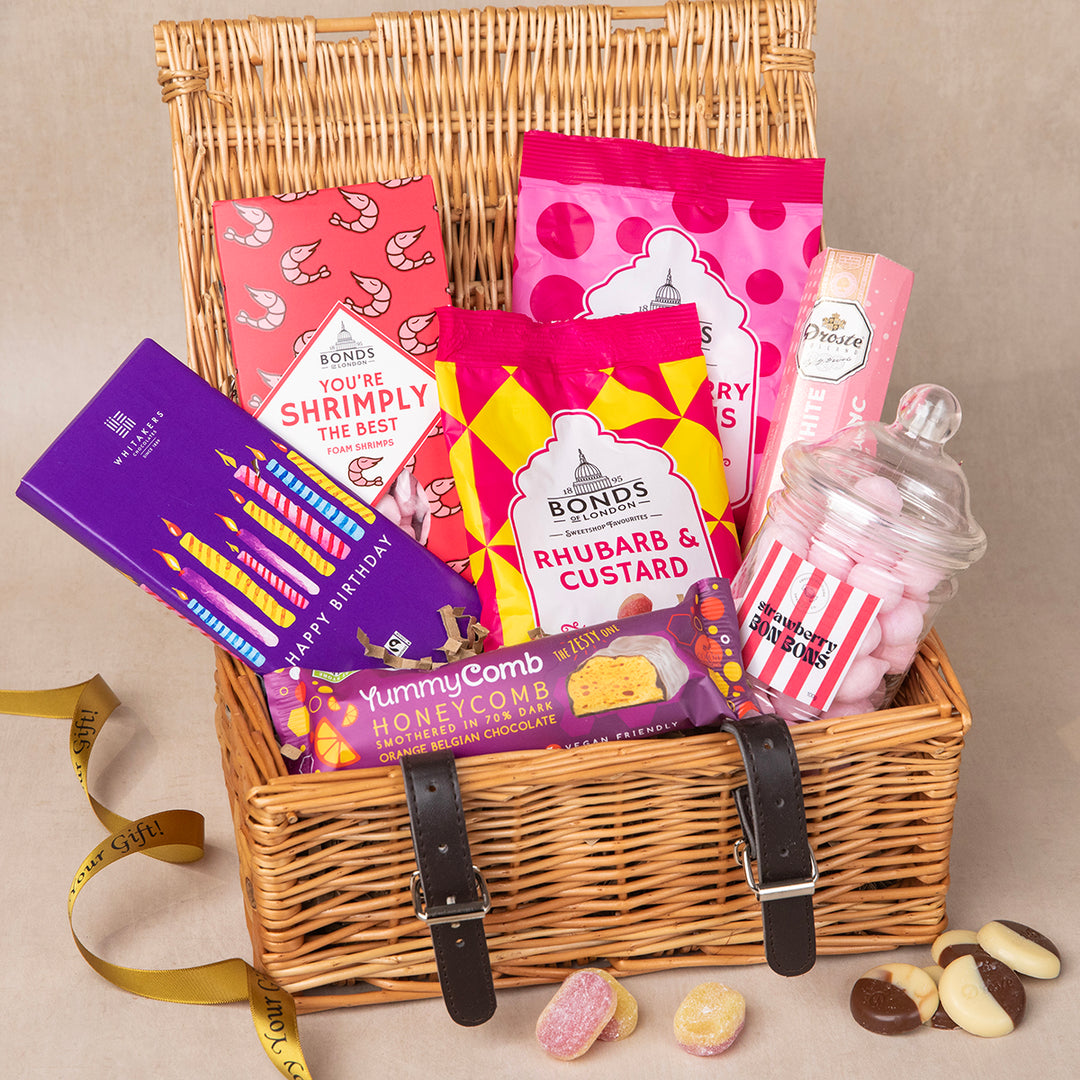 A wicker hamper gift basket with straps, filled with an assortment of sweet treats including chocolate bars, gummy candies, and a jar of bon bons. The basket is presented on a neutral background.